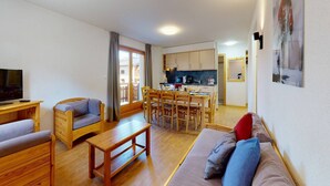 A cozy living room with a sofa, a wooden table, and a TV. There's a kitchen area with cabinets and a dining table. - Modern apartment with balcony (Haute-Nendaz)