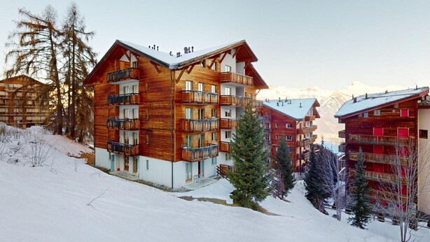 A multi-story wooden building with balconies, surrounded by snow-covered terrain and mountains in the background. - Lovely apartment with valley view (Haute-Nendaz)