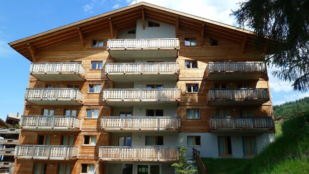 Un immeuble en bois à plusieurs étages, doté de balcons, d’une entrée centrale et entouré d’une zone verdoyante.