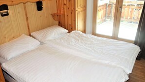 Un lit double avec du linge de lit blanc, dans une pièce aux murs en bois et dotée d’une porte-fenêtre coulissante donnant sur l’extérieur.