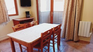 Een eethoek met een houten tafel en stoelen, een koffiezetapparaat, een televisie aan de muur en een glazen schuifdeur naar buiten.