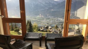 A room with a large window offering a view of a town and mountains. There are two black chairs in the room. - Mountain apartment with parking (Veysonnaz)
