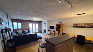 A modern living room with a sofa, a dining area with a table and chairs, wooden flooring, and a kitchen area with a counter and cabinets. - Mountain apartment with parking (Veysonnaz)