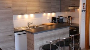 A modern kitchen with wooden cabinets, a stainless steel sink, a coffee machine, and a small island with bar stools. - Mountain apartment with parking (Veysonnaz)