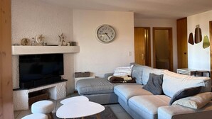 Un salon avec un téléviseur à écran plat, un canapé gris, une table basse blanche et une horloge au mur.