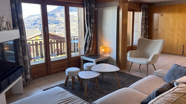 Un salon chaleureux avec un canapé, un fauteuil, une petite table et un téléviseur. De grandes fenêtres offrent une vue sur une terrasse en bois et les montagnes.
