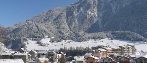 A snow-covered mountain village with traditional chalet-style buildings, a ski slope, and a clear blue sky.
