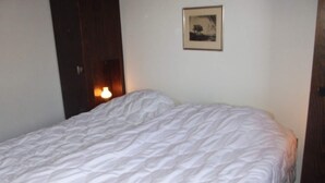 A bedroom with a large bed, a framed picture on the wall, and a door with a light on the left side. - Pleasant apartment with balcony (Veysonnaz)