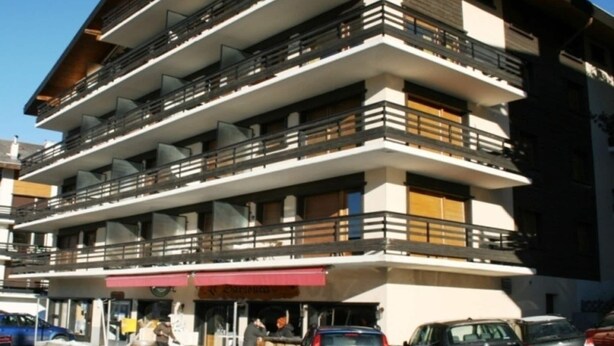 A multi-story building with balconies, a ground-level entrance, and parked cars. - Pleasant apartment with balcony (Veysonnaz)