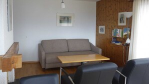 A cozy living room with a sofa, a wooden table, and a bookshelf with books and a lamp. - Pleasant apartment with balcony (Veysonnaz)