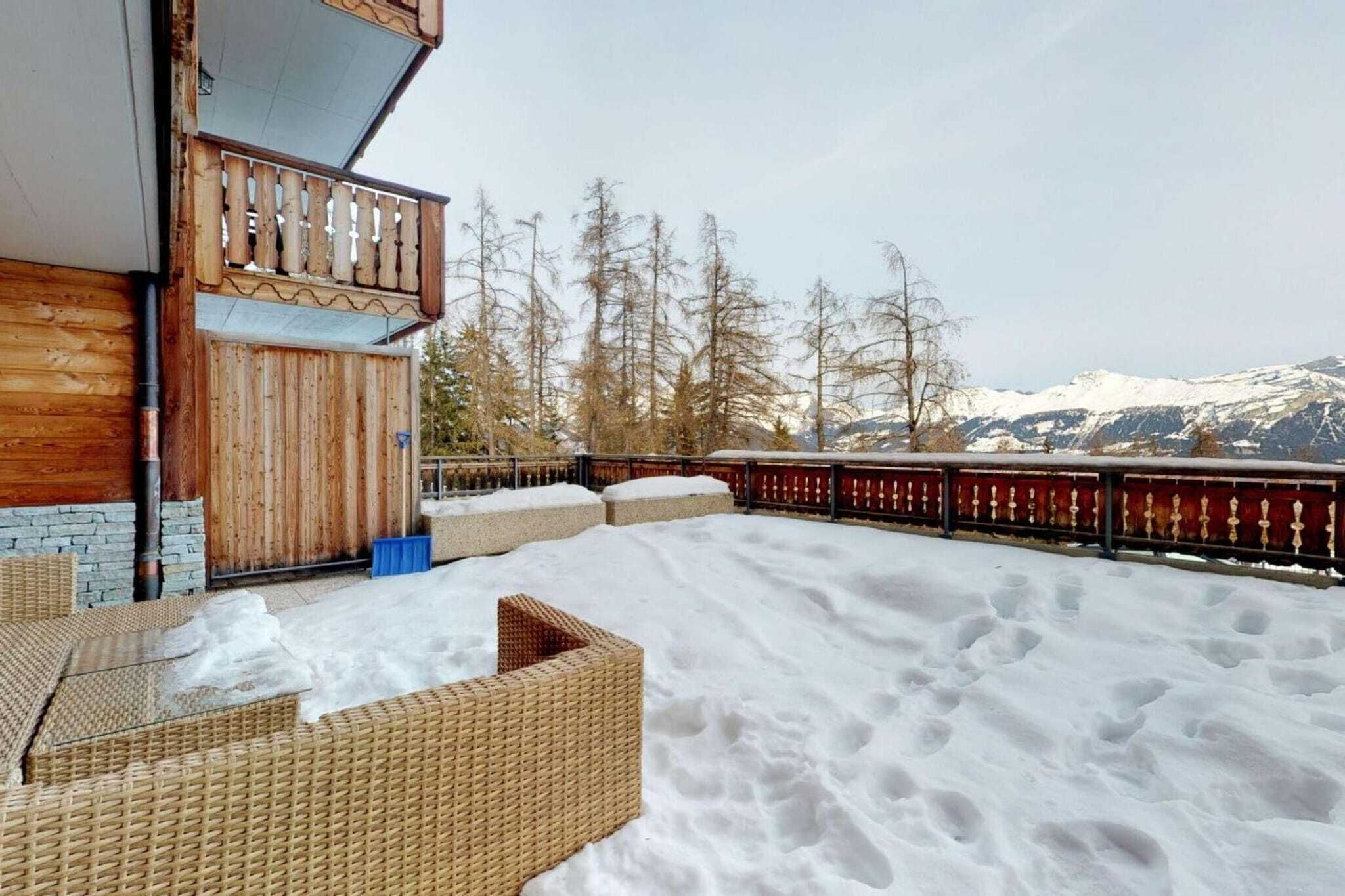Ett trädäck med möbler av rotting, ett rött räcke och ett snötäckt berg i bakgrunden.