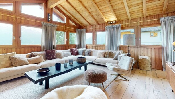 A spacious living room with wooden walls and ceiling, large windows, a sofa set, a coffee table, and a lamp. - Beautiful chalet in Le Bouveret with garden (Les Masses)