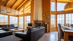 Ein geräumiges Wohnzimmer mit Kamin, Ledersofas, einem Essbereich mit Holztisch und Stühlen sowie großen Fenstern mit Blick auf die Berge.