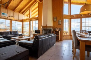 Un vaste salon doté d’une cheminée, de canapés en cuir, d’un coin repas avec une table et des chaises en bois, et de grandes fenêtres offrant une vue sur les montagnes.