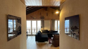 A spacious living area with a large sofa, a brown armchair, and a dining table. There are large windows with curtains, a wooden ceiling, and artwork on the walls. - Family apartment with terrace (Veysonnaz)