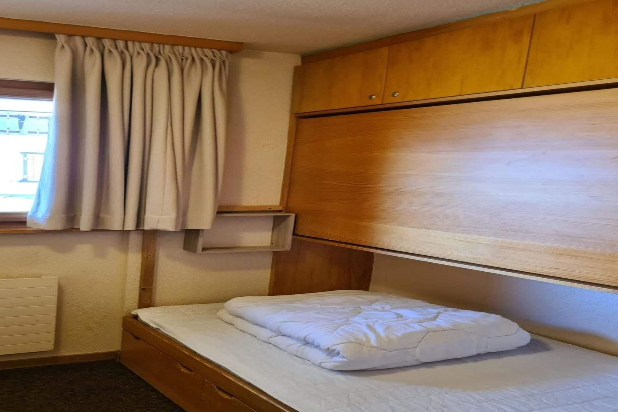 Une petite chambre avec un lit simple, une tête de lit en bois, une fenêtre avec des rideaux et une étagère intégrée au-dessus du lit.
