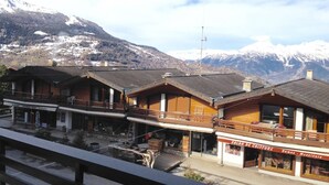 Une rangée de chalets de montagne en bois, dotés de balcons, un salon de coiffure bien visible, et des montagnes enneigées en arrière-plan.