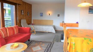 Une pièce avec un canapé rouge, un lit simple, une table recouverte d’une nappe jaune et une chaise.