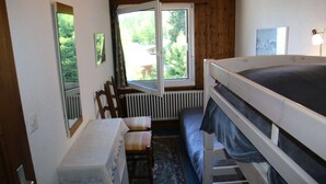 A small, cozy room with a bunk bed, a wooden chair, a small table, and a window with a view of greenery outside.