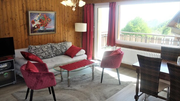 A living room with a sofa, red chairs, a glass coffee table, a painting, and a view of a balcony with a railing.