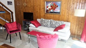 A living room with a white sofa, red and patterned cushions, a pink chair, a wooden TV stand, and a painting on the wall.