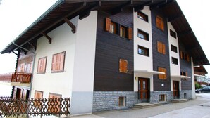A multi-story building with a combination of white and dark wooden exterior walls, multiple windows with wooden shutters, and a balcony on the ground floor.