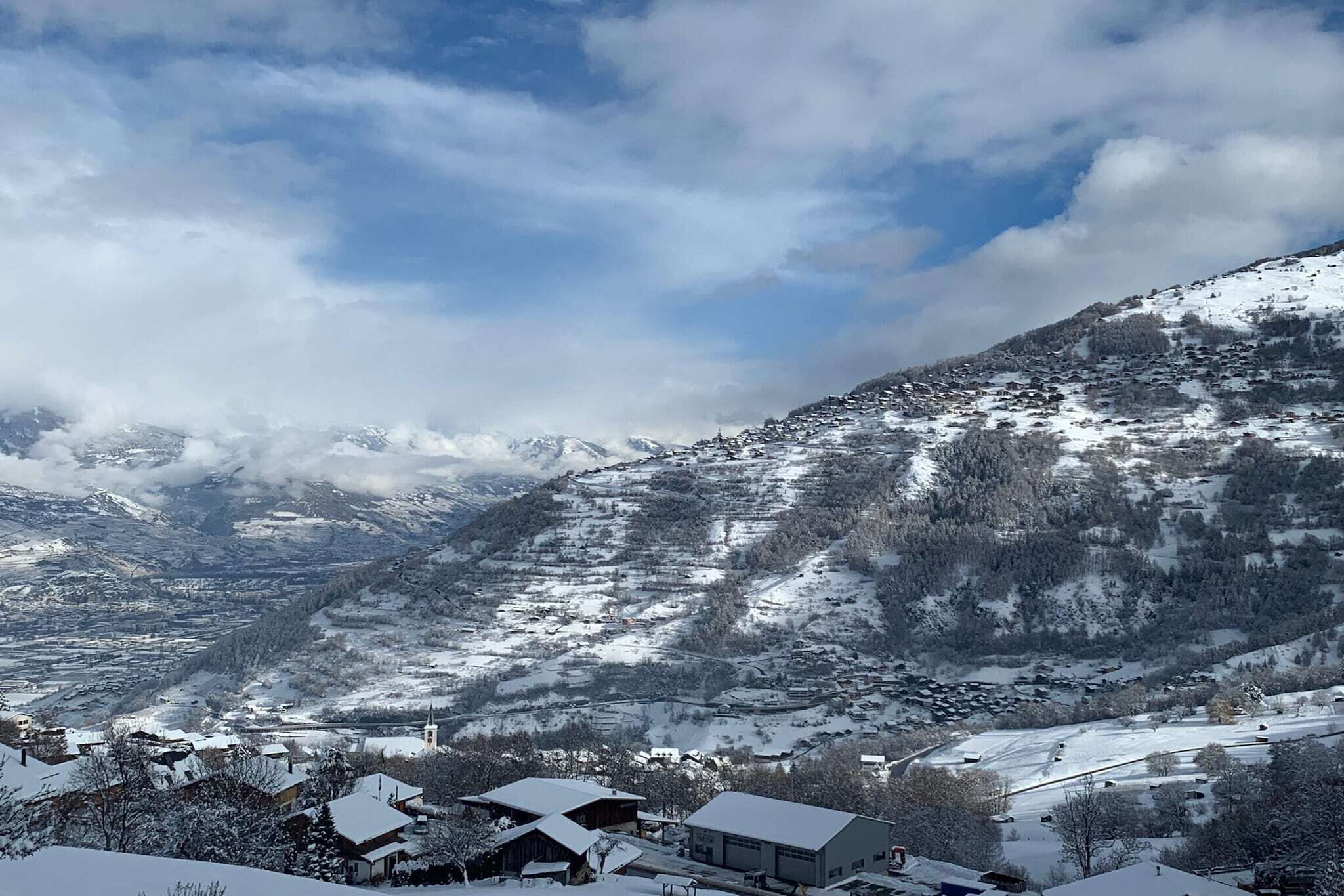 Un village de montagne enneigé, avec des maisons et une église, entouré de collines couvertes de neige et d’un ciel partiellement nuageux.