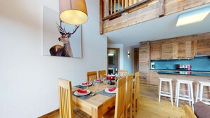 Un comedor con una mesa de madera dispuesta para seis personas, un cuadro de un ciervo y una cocina con armarios y taburetes de madera.