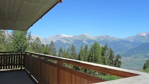 Un balcon doté d’une balustrade en bois, d’où l’on peut admirer un paysage montagneux avec des sommets enneigés et un ciel bleu dégagé.