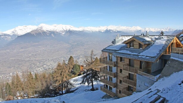 A multi-story hotel with a snow-covered roof, wooden balconies, and a view of snow-capped mountains. - Modern apartment with sauna (Veysonnaz)
