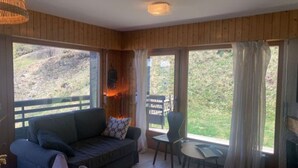 A cozy room with a wooden interior, a sofa, a small table, and a view of a grassy landscape through large windows. - Apartment with Rhone valley view (Veysonnaz)