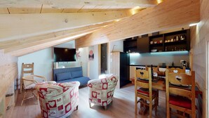 A cozy loft-style room with a dining area, wooden beams, and a kitchen in the background.