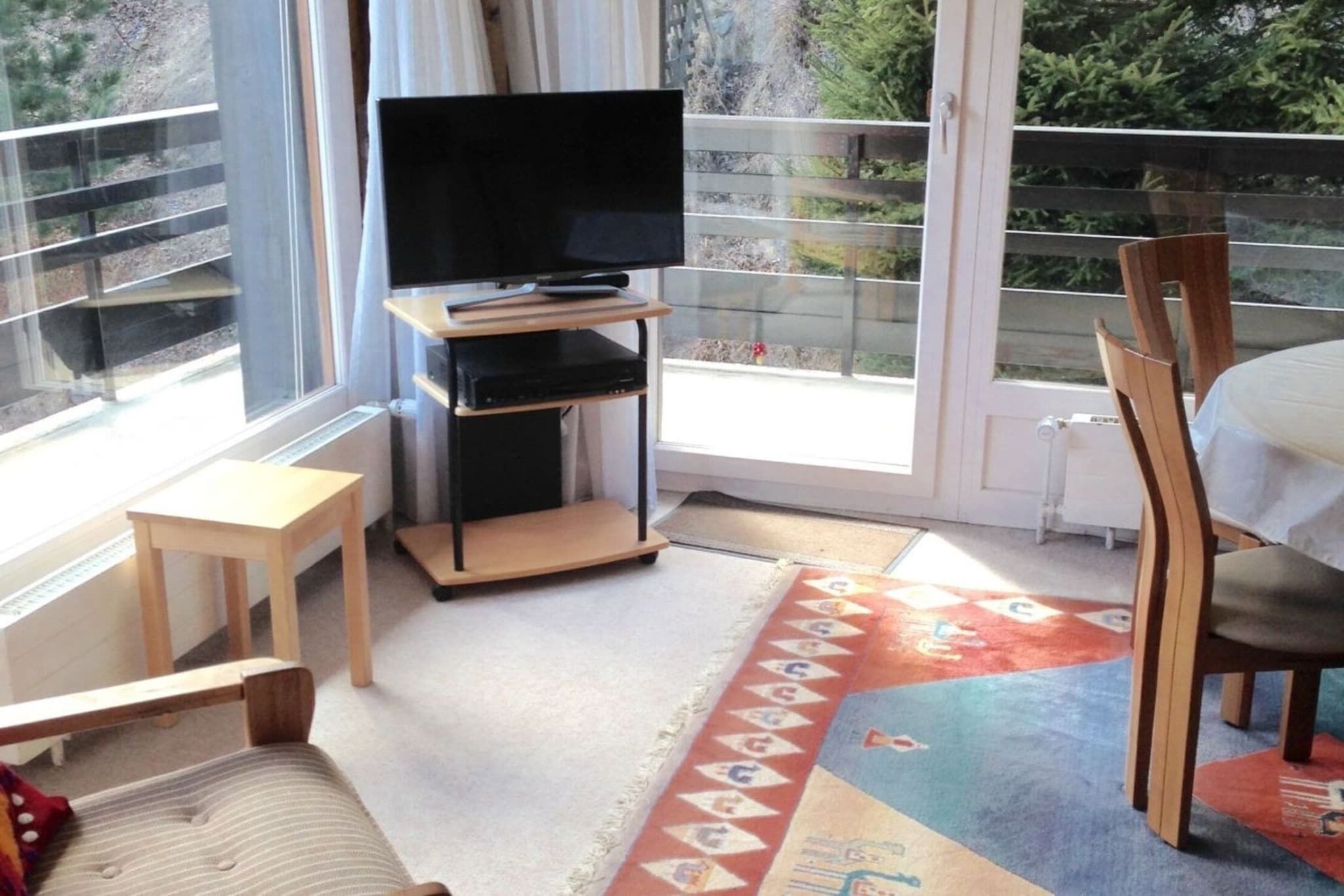 Una habitación con una televisión sobre un mueble bajo, una mesita, una silla y una alfombra colorida.