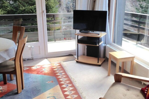 Una habitación con un ventanal grande, un televisor sobre un mueble bajo, una mesa de comedor con sillas y un taburete de madera pequeño.