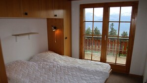 Ein Schlafzimmer mit Bett, Holz-Kopfteil, Regal, Wandleuchte und einer Glas-Schiebetür mit Blick auf die Berge.