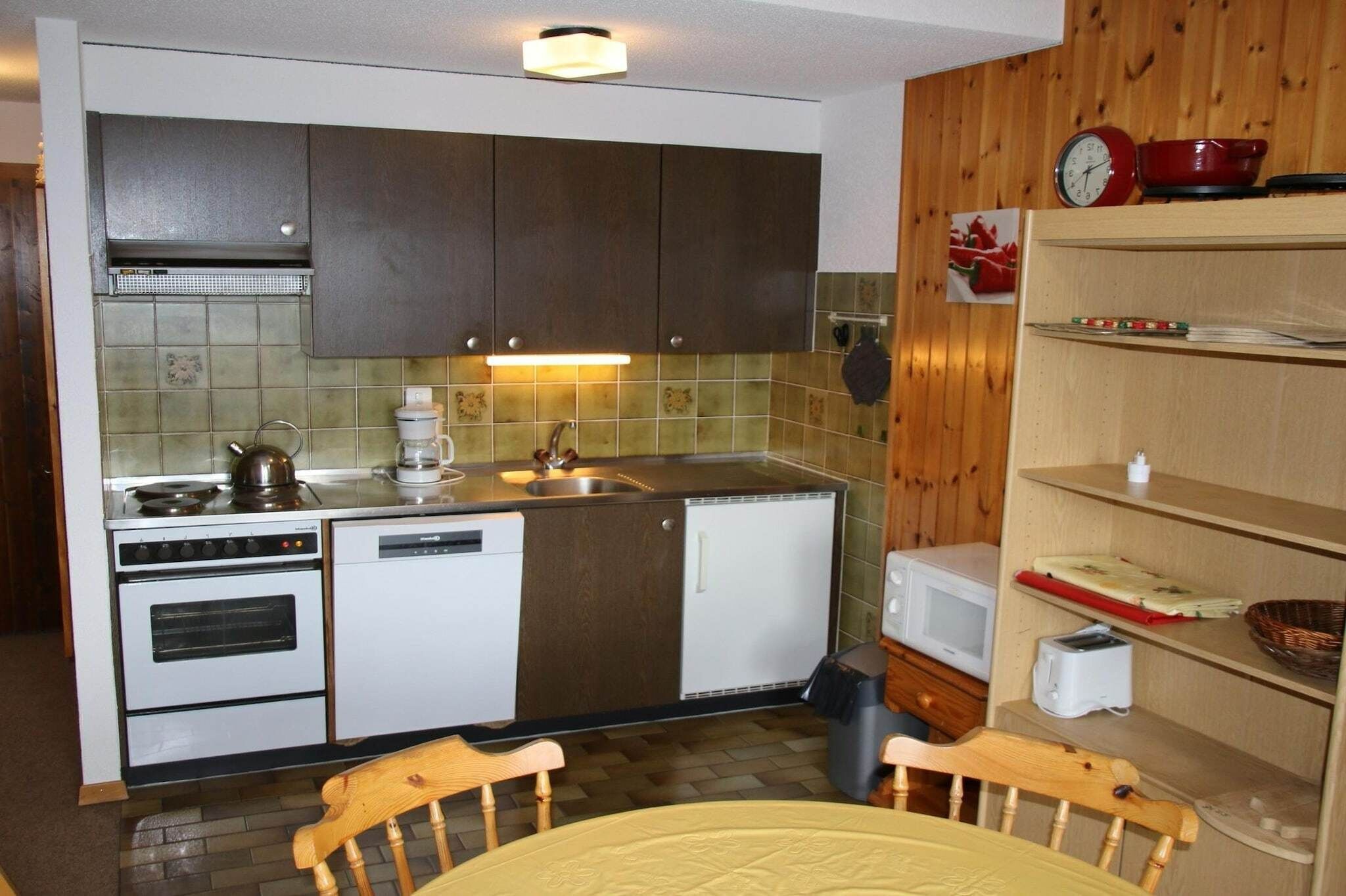 Une petite cuisine équipée d’une cuisinière, d’un évier et d’un four à micro-ondes. On aperçoit une chaise en bois et une table au premier plan.
