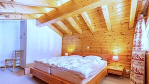 Une chambre à coucher en bois, avec un lit, une table de chevet, une lampe, une chaise et une fenêtre avec un rideau.