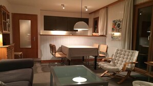 Ein modernes Wohnzimmer mit einem Glastisch, einer Couch, einem Esstisch mit Stühlen und einem großen Fenster mit Vorhängen.