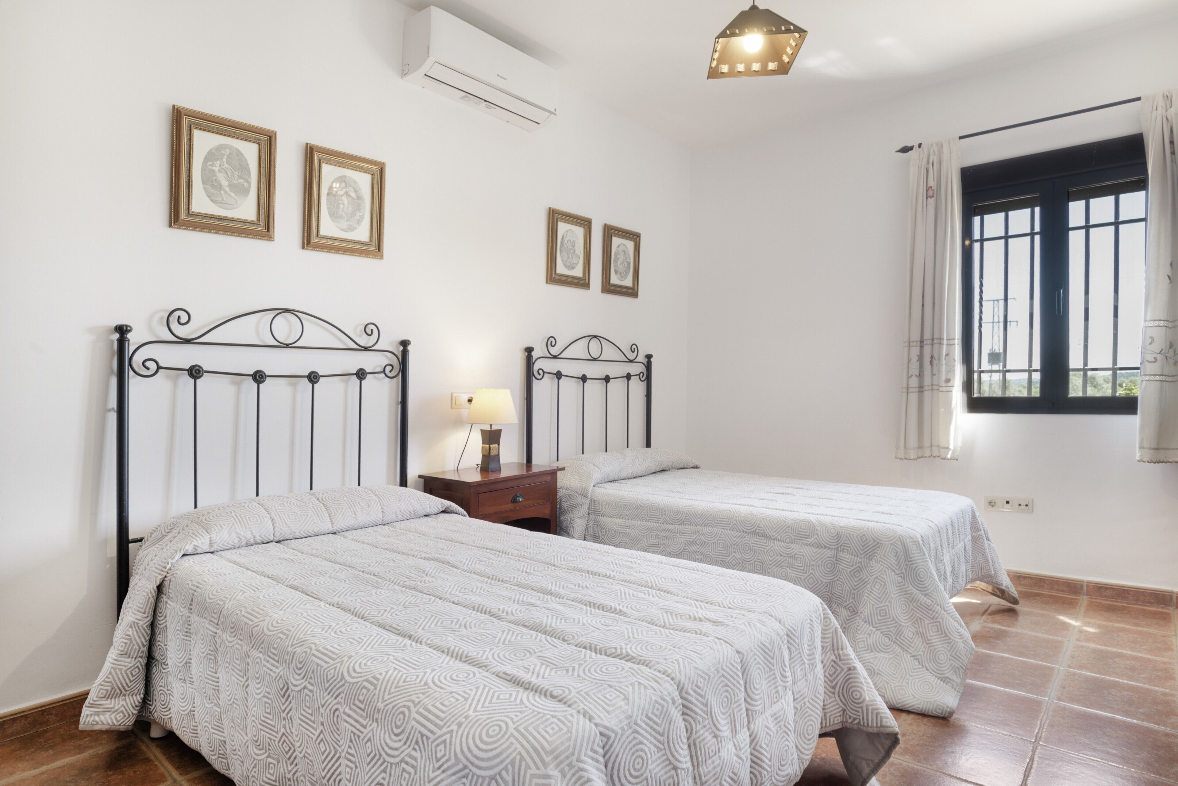 Une chambre avec deux lits, une table de chevet avec une lampe, des tableaux encadrés au mur et une fenêtre avec des rideaux.
