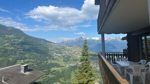 A balcony with a table and chairs overlooking a mountainous landscape with a village below and snow-capped peaks in the distance.