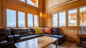 Een houten chalet met een leren bank, een houten bijzettafeltje en grote ramen met uitzicht op de bergen.