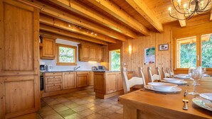 Een houten interieur met een keuken en eethoek, met houten keukenkastjes, een betegelde vloer en een eettafel gedekt voor vier personen.