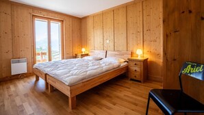 Slaapkamer met houten lambrisering, een bed, bedlampjes, een stoel en uitzicht op bergen door een groot raam.