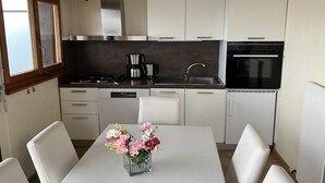 Une cuisine moderne avec des armoires blanches, une hotte de cuisinière en acier inoxydable, une machine à café et un évier. On y trouve une table à manger avec des chaises et un vase de fleurs.