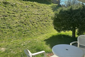 Une terrasse avec une table et des chaises blanches, entourée d’une pelouse et d’un petit arbre.