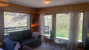 A cozy room with wooden walls, a sofa, a table, and large windows offering a view of a grassy landscape. - Apartment in Veysonnaz near Ski area (Veysonnaz)