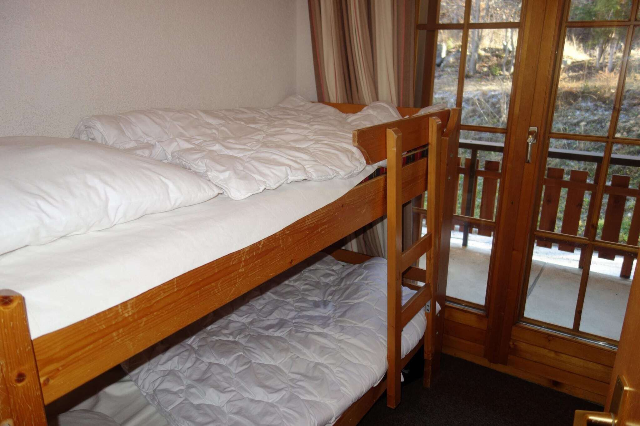 A bunk bed with white bedding and a wooden headboard, a window with curtains, and a view of a wooden deck outside.