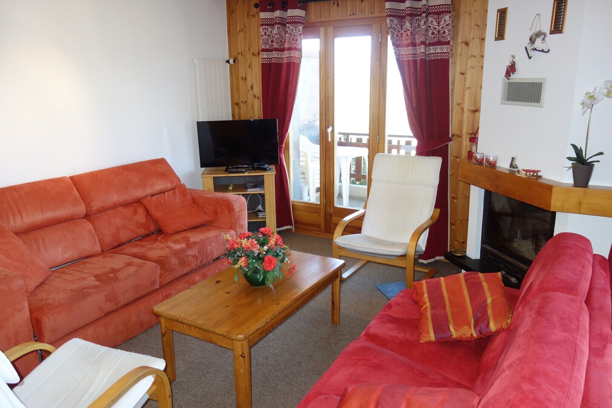 A living room with a red sofa, a white armchair, a wooden coffee table with a flower vase, a television, and a fireplace.