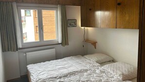 A bedroom with a bed, wooden wardrobe, a window with curtains, a radiator, and a small shelf. - Charming apartment in Vaysonnaz (Veysonnaz)