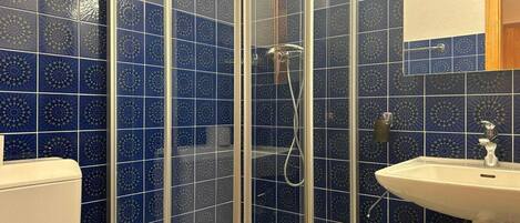 Ein Badezimmer mit blauen, gemusterten Fliesen, einer Glasdusche, einem weißen Waschbecken und einer Holztür.